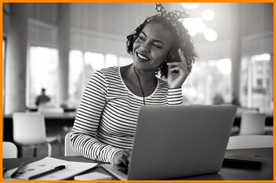 Dynamic young female university student listening to music on headphones working online on laptop sitting at table